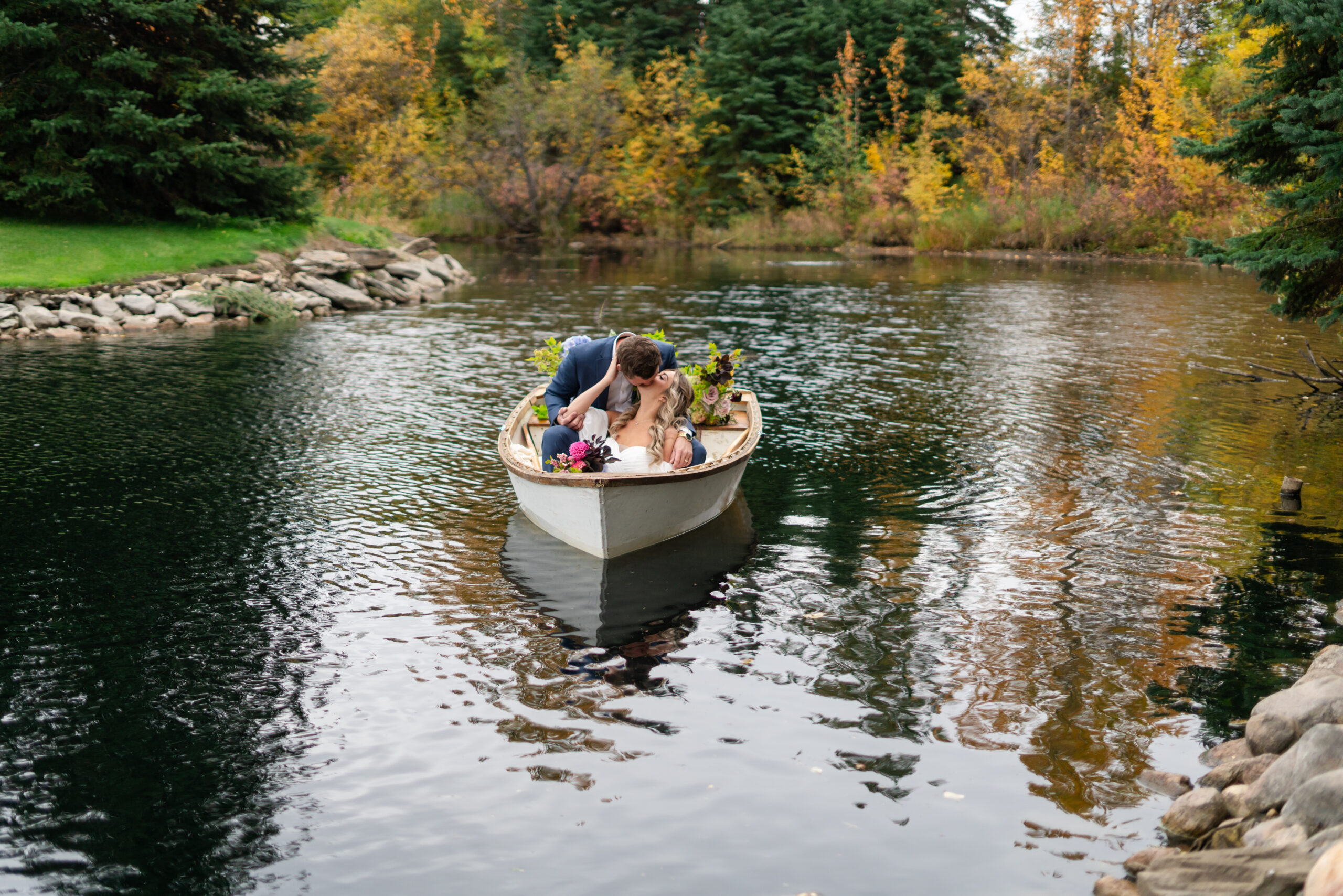 A bride and groom in a row boat on a pond surronded by fall foliage. the couple is kissing